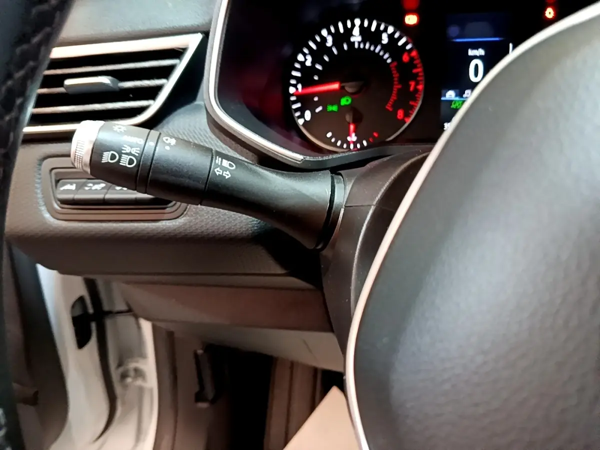 Gros plan sur le commodo des phares et clignotants à gauche du volant noir dans l’habitacle d’une Renault Clio blanche.