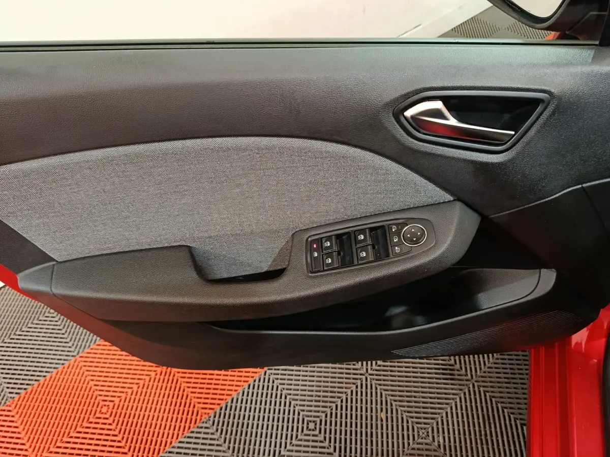 Vue intérieure côté conducteur de la porte d’une Renault Clio rouge 2024 avec commandes électriques et garniture tissu grise.