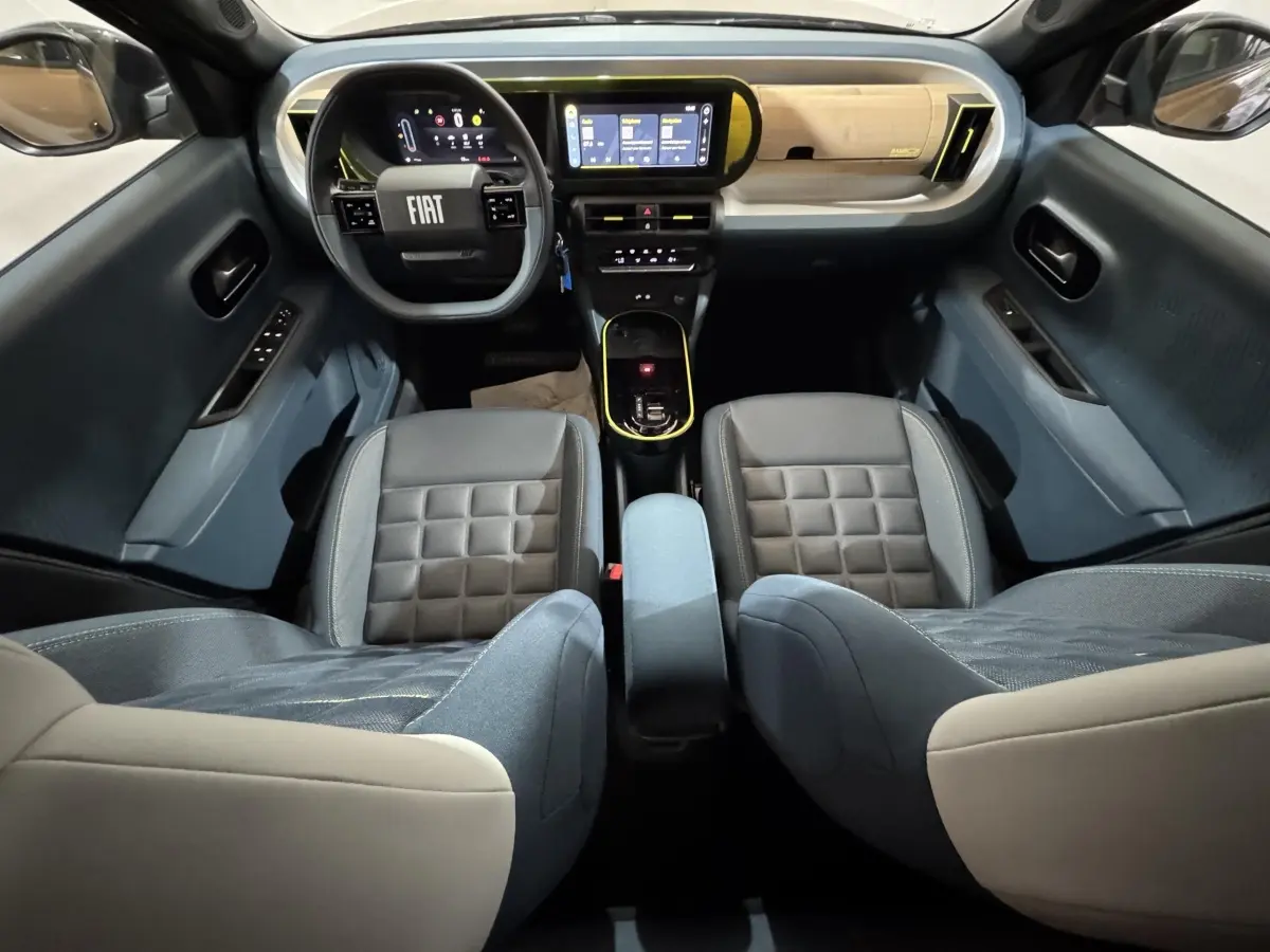 Vue intérieure avant de la Fiat Grande Panda 2025, montrant tableau de bord marron clair et sièges bi-ton gris et beige.
