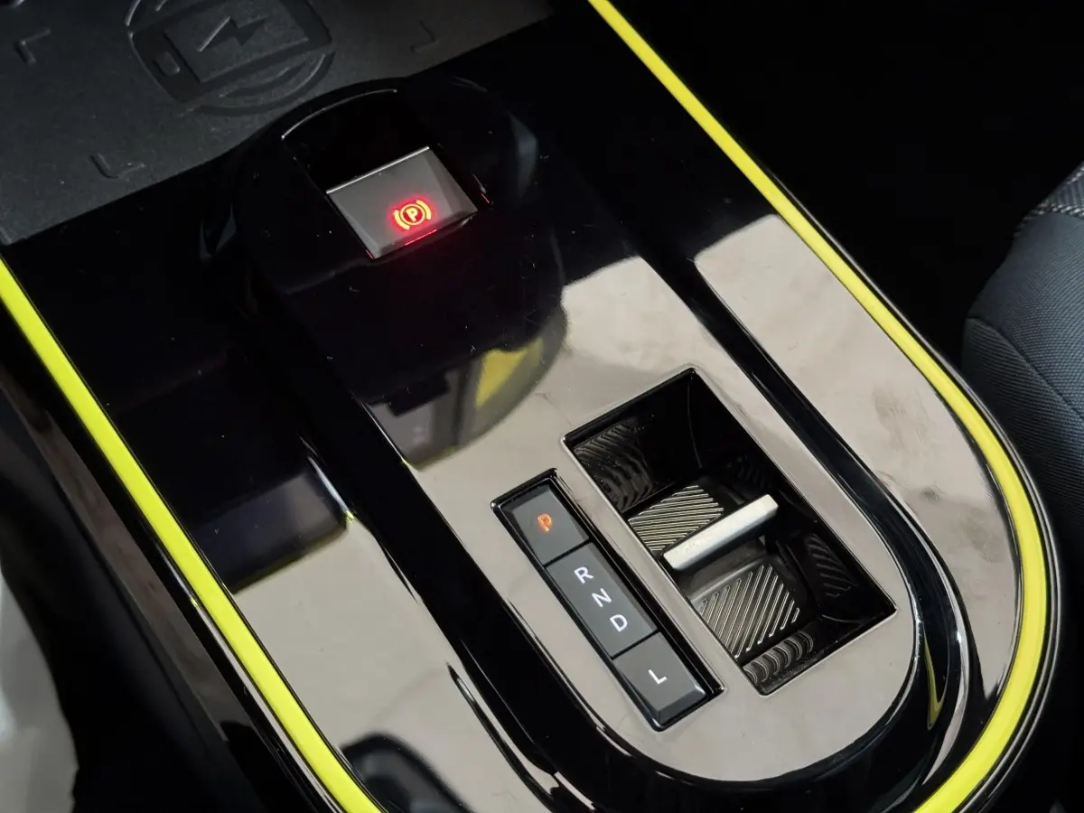 Gros plan sur la console centrale noire brillante avec bouton frein électrique rouge et sélecteur de boîte auto entouré de jaune.