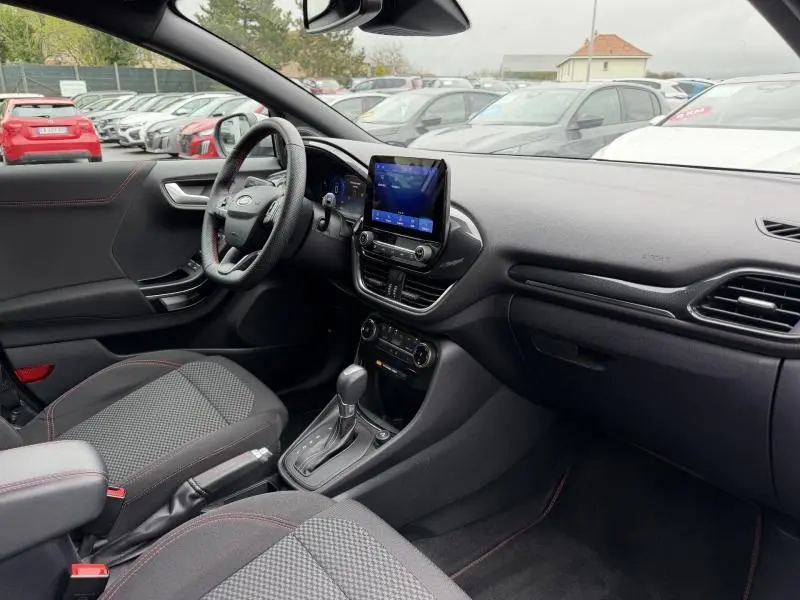Vue intérieure côté conducteur du Ford Puma 2024, volant sport, écran tactile et sièges tissu noir avec surpiqûres rouges.
