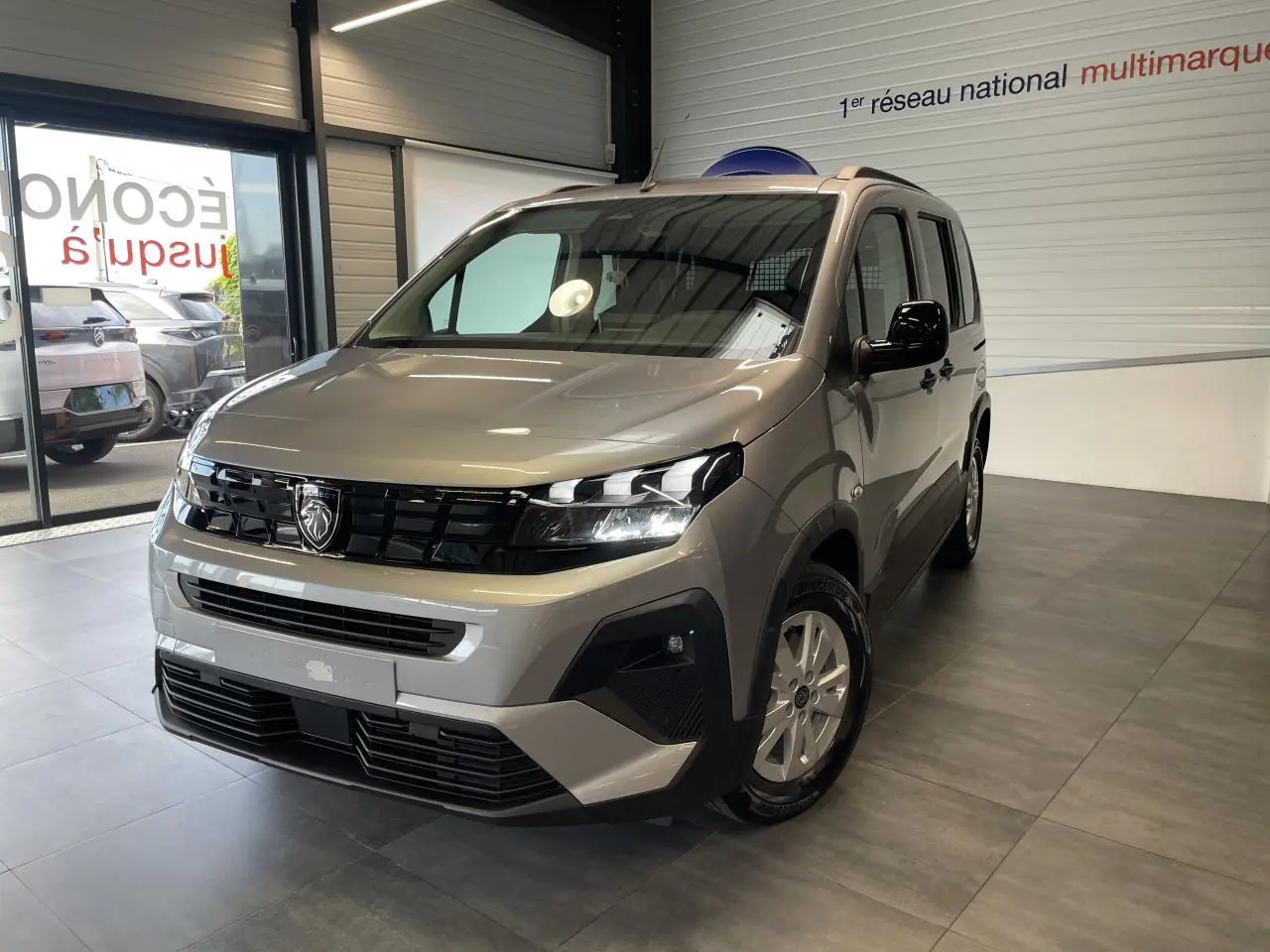 Peugeot Rifter gris Artense vu en 3/4 avant droit dans un showroom, avec calandre noire et jantes tôles 16 pouces.