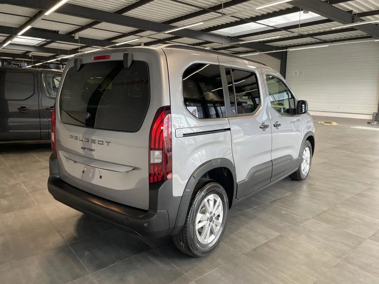 Vue 3/4 arrière droit du Peugeot Rifter gris Artense avec feux arrière verticaux et jantes tôles 16 pouces en showroom.