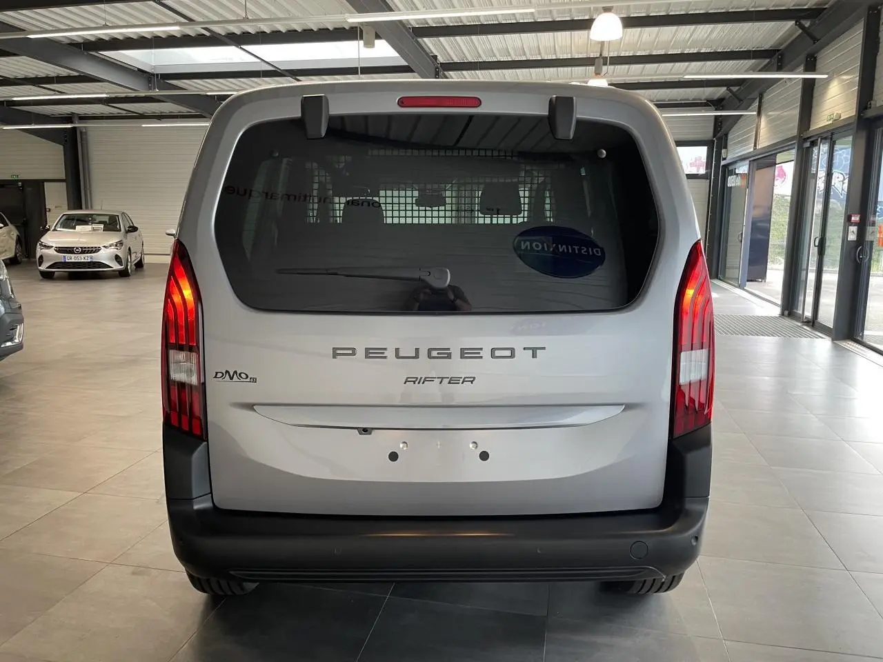 Vue arrière d’un Peugeot Rifter gris Artense avec hayon vitré et feux arrière verticalisés en intérieur showroom.