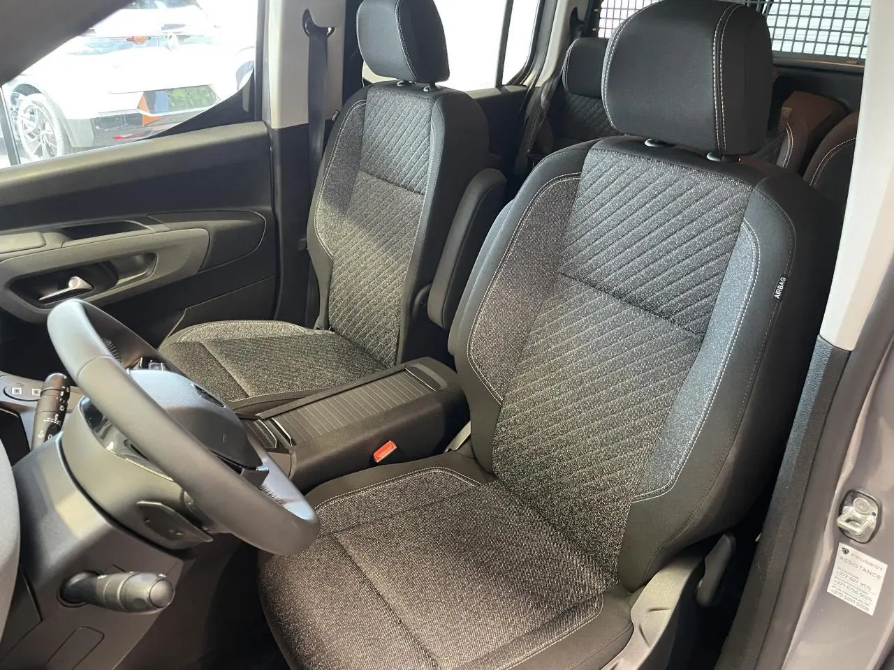 Vue intérieure sur les sièges avant en tissu gris du Peugeot Rifter 2026, avec accoudoirs et volant multifonction cuir.