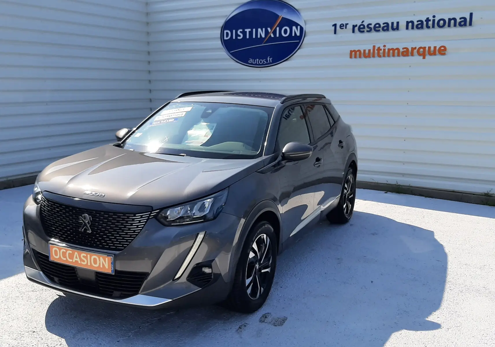 Peugeot 2008 gris Artense vue 3/4 avant droit, avec calandre noire et feux LED distinctifs, stationnée devant un mur blanc.
