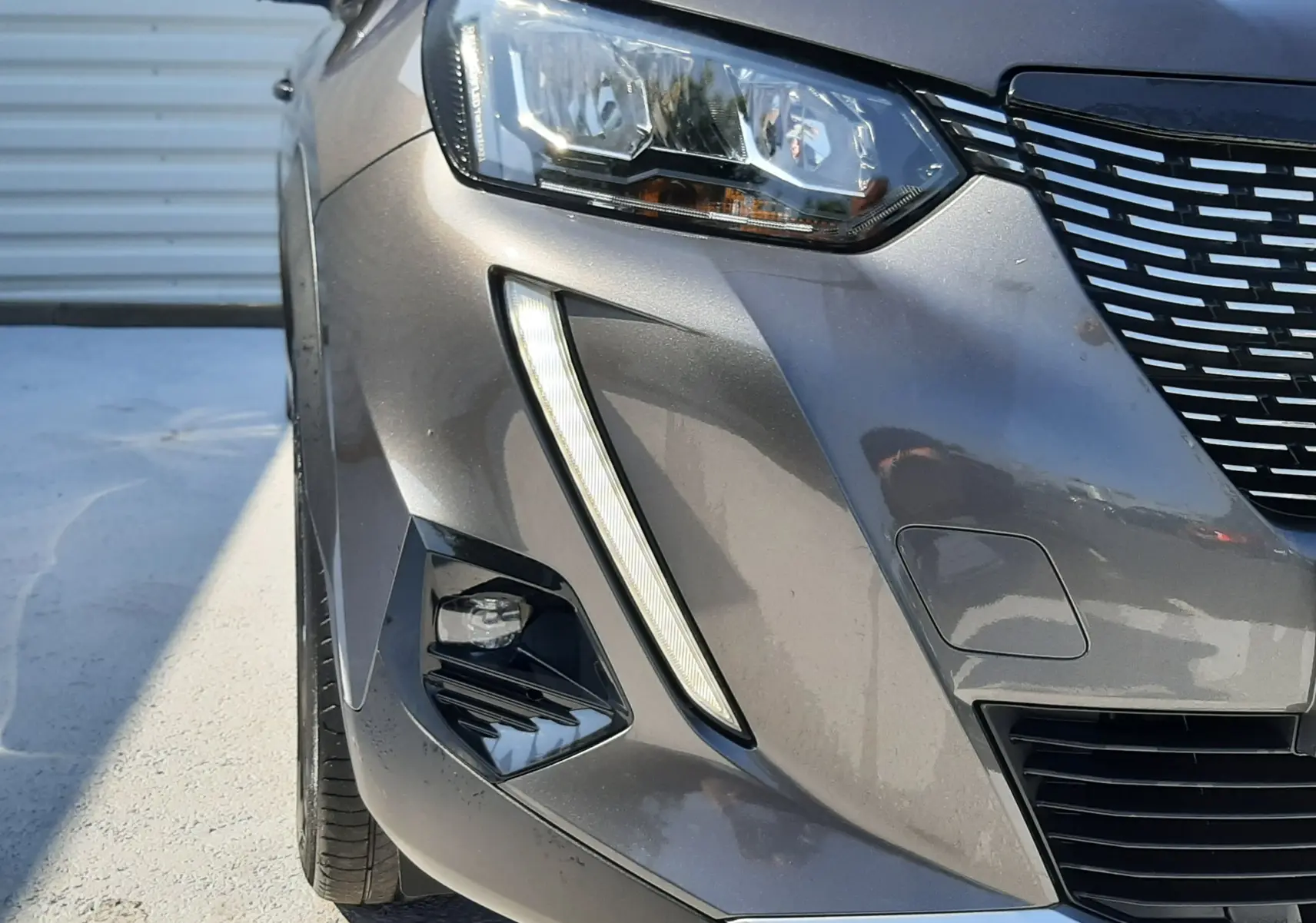 Gros plan sur l'avant gauche d'une Peugeot 2008 gris Artense, mettant en valeur le phare et la calandre distinctive.