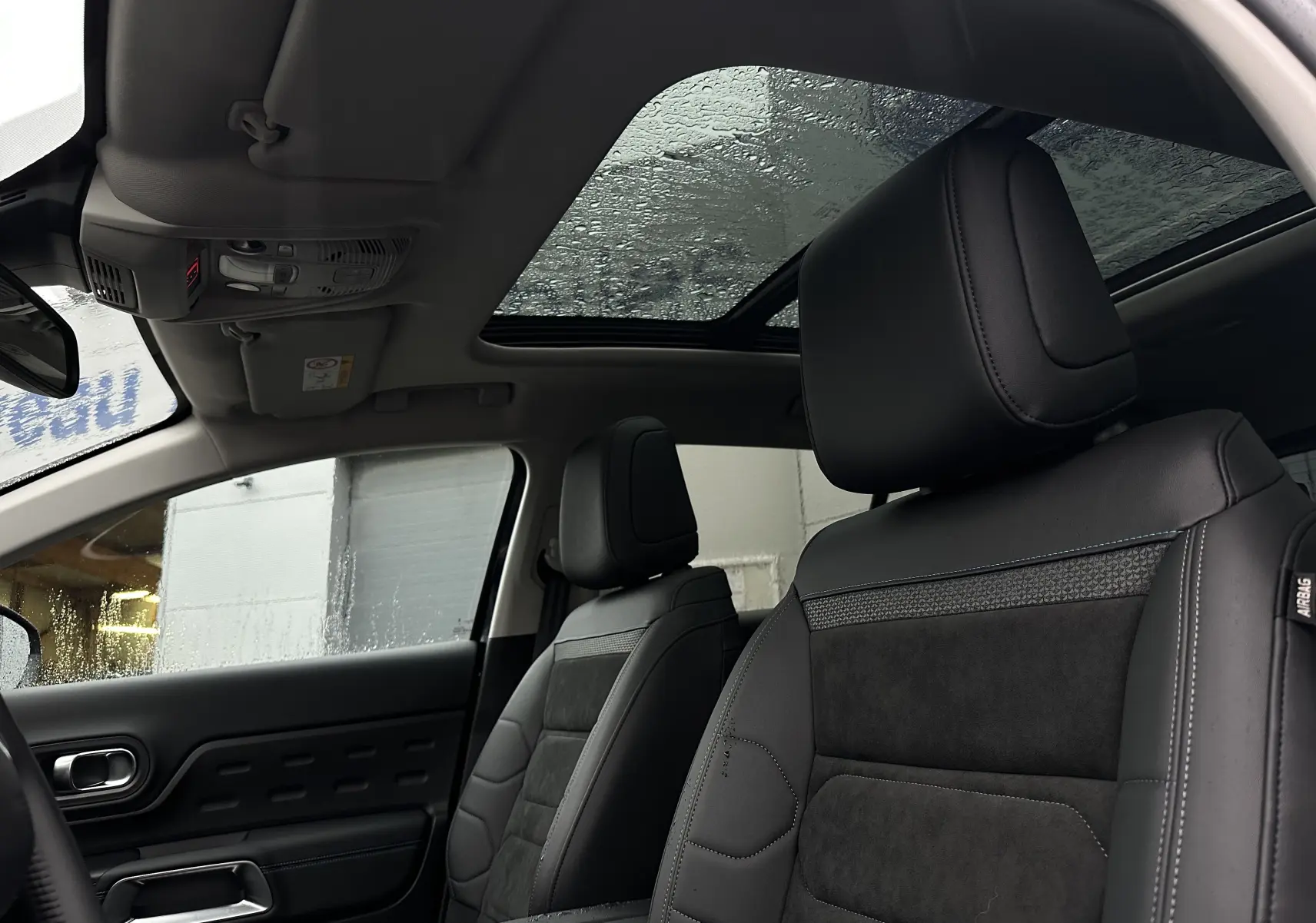 Vue intérieure avant du Citroën C5 Aircross 2025, sièges cuir noir et toit ouvrant panoramique vitré sous la pluie.