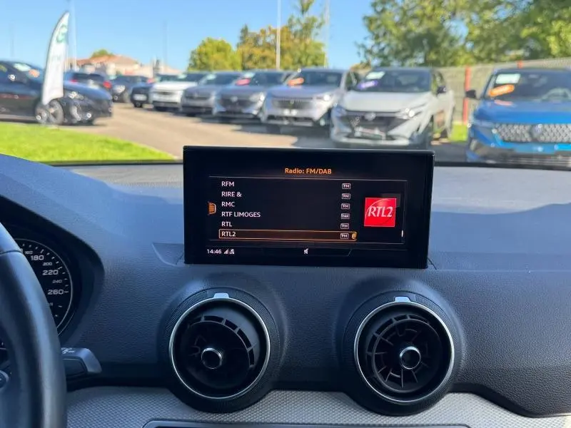 Vue intérieure du tableau de bord de l'Audi Q2 2023, écran central affichant la radio FM/DAB avec ventilation circulaire noire.