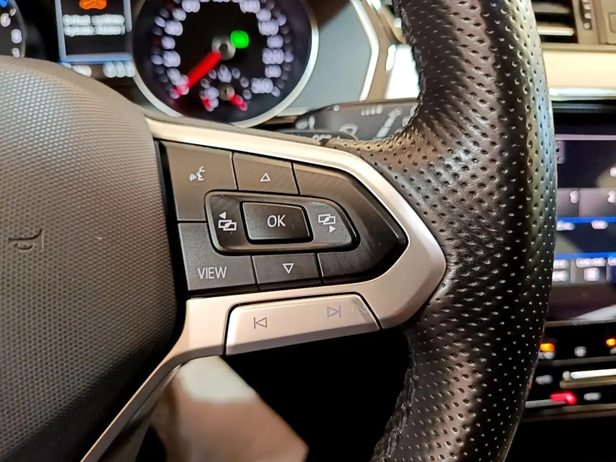 Gros plan sur les commandes gauche du volant en cuir perforé de la Volkswagen Passat hybride blanche, avec tableau de bord digital en arrière-plan.