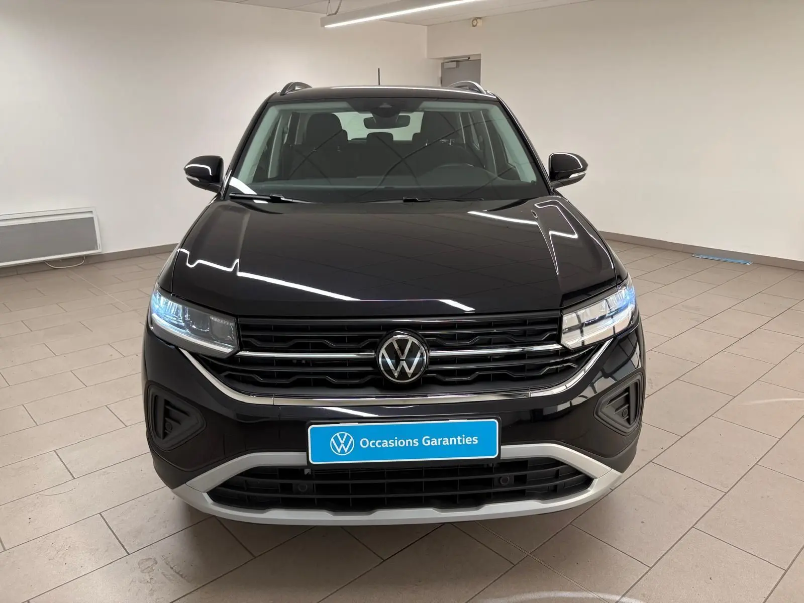 Vue frontale du Volkswagen T-Cross noir intense 2024 avec calandre chromée et phares LED allumés dans un showroom.