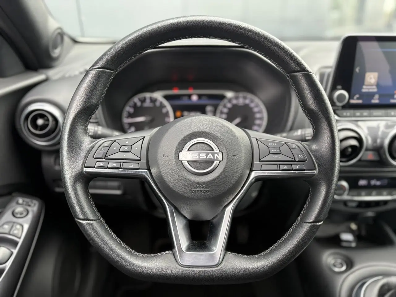 Vue rapprochée du volant en cuir noir du Nissan Juke blanc perle 2024, avec commandes intégrées et tableau de bord flou.