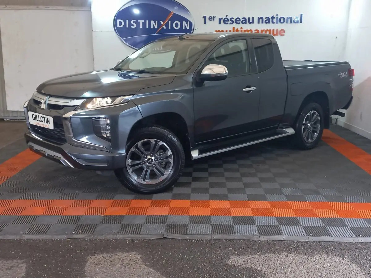Mitsubishi L200 gris foncé en 3/4 avant droit, avec benne pick-up et jantes alliage visibles en intérieur showroom.