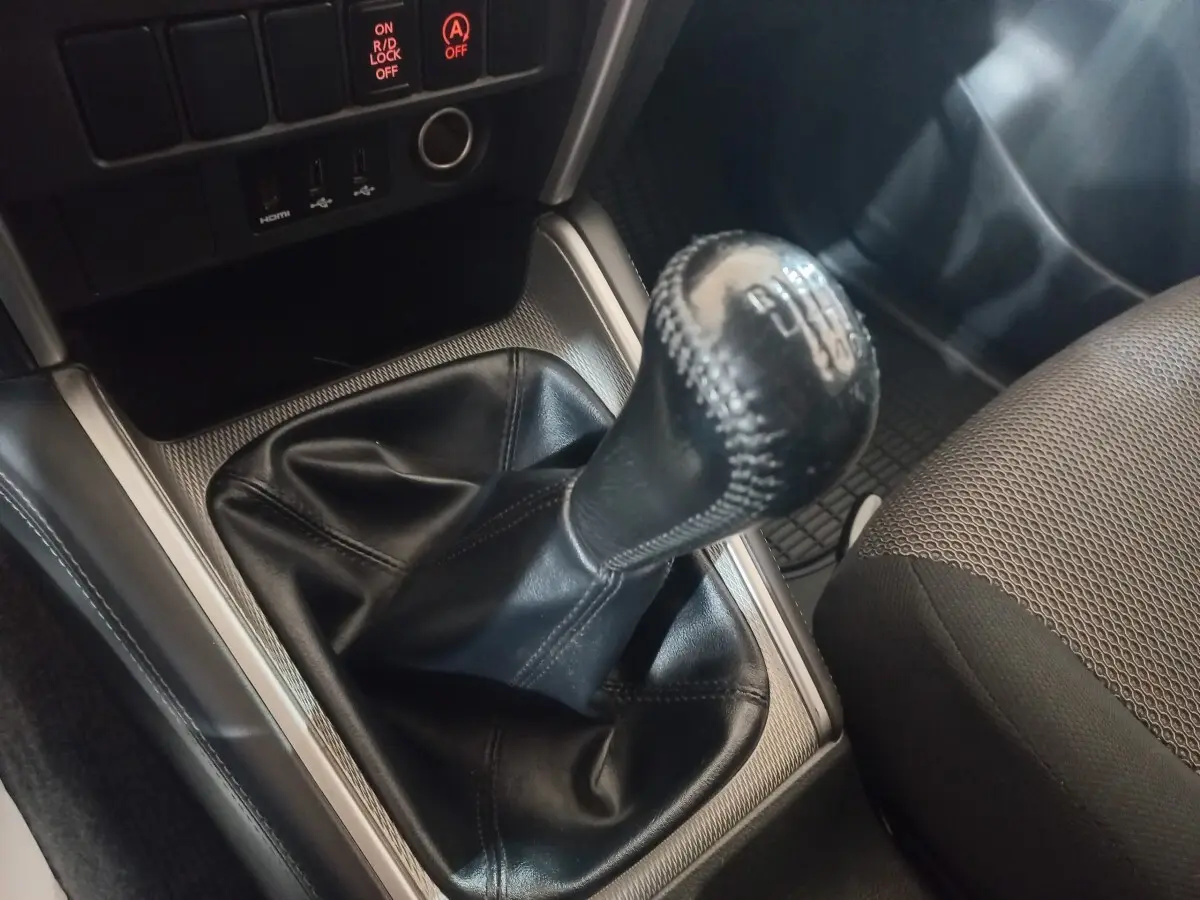 Gros plan sur le levier de vitesses manuel en cuir noir du Mitsubishi L200, avec console argentée et siège tissu gris visible.