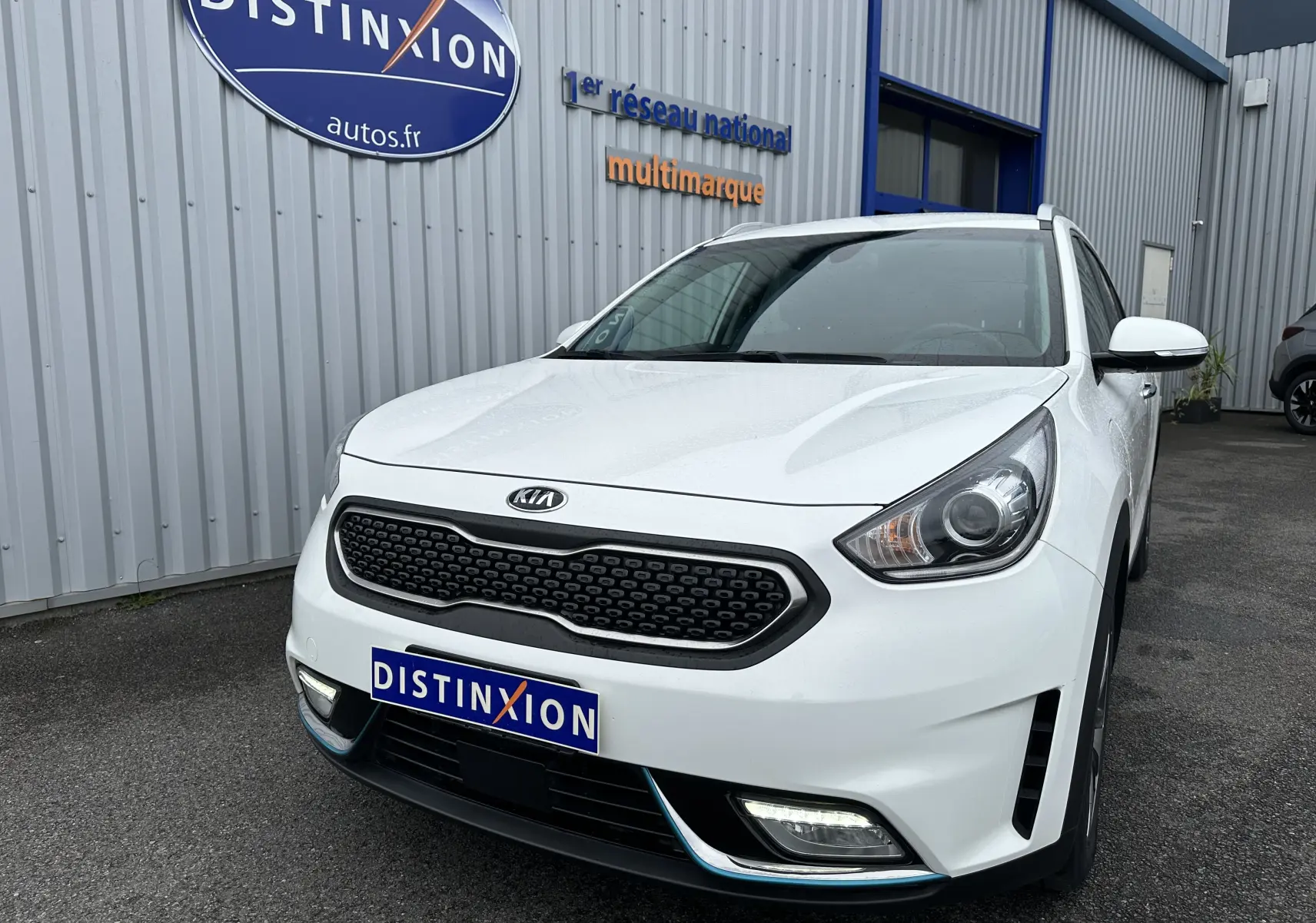 Vue 3/4 avant d'un Kia Niro hybride rechargeable blanc céleste, avec calandre noire et feux LED visibles, stationné devant un bâtiment.