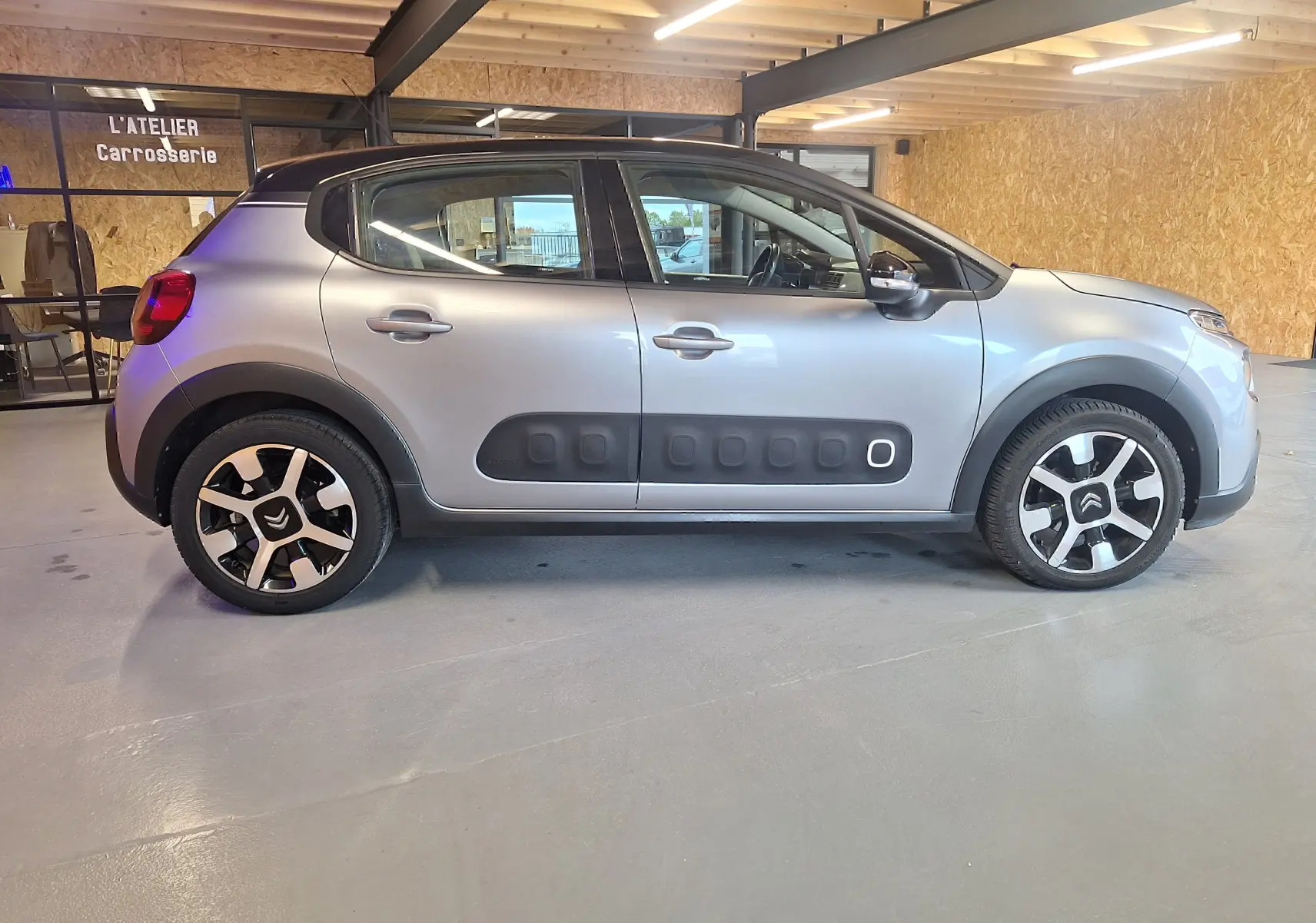 Profil droit d'une Citroën C3 gris clair avec protections latérales noires et jantes aluminium stylisées dans un atelier carrosserie.