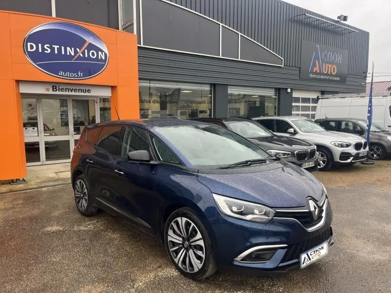 Renault Grand Scenic bleu Cosmos en 3/4 avant droit, garé devant un concessionnaire sous la pluie.