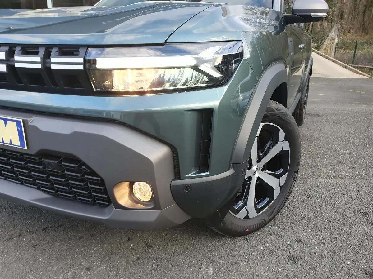 Gros plan sur l'avant droit du Dacia Duster vert cèdre, mettant en valeur le phare allumé et la jante alliage 18 pouces.