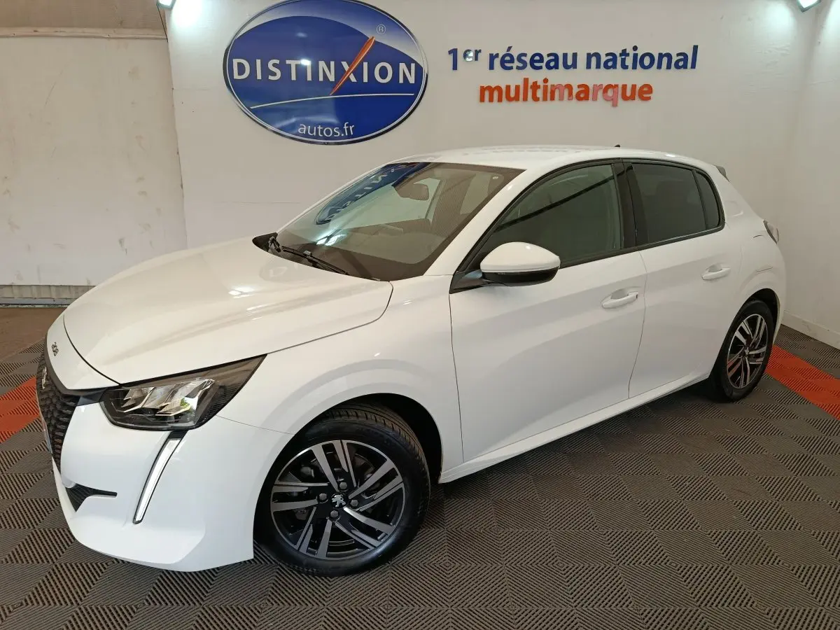 Peugeot 208 blanc vue 3/4 avant droit en intérieur, avec jantes alliage noires et feux LED distinctifs.