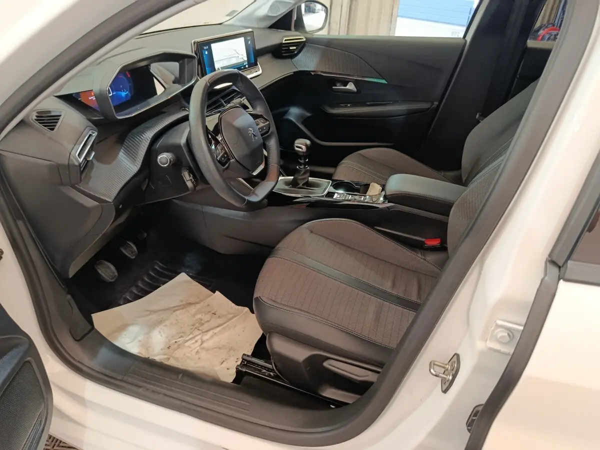 Vue intérieure côté conducteur de la Peugeot 208 blanche, montrant le tableau de bord numérique et la console centrale avec boîte manuelle.