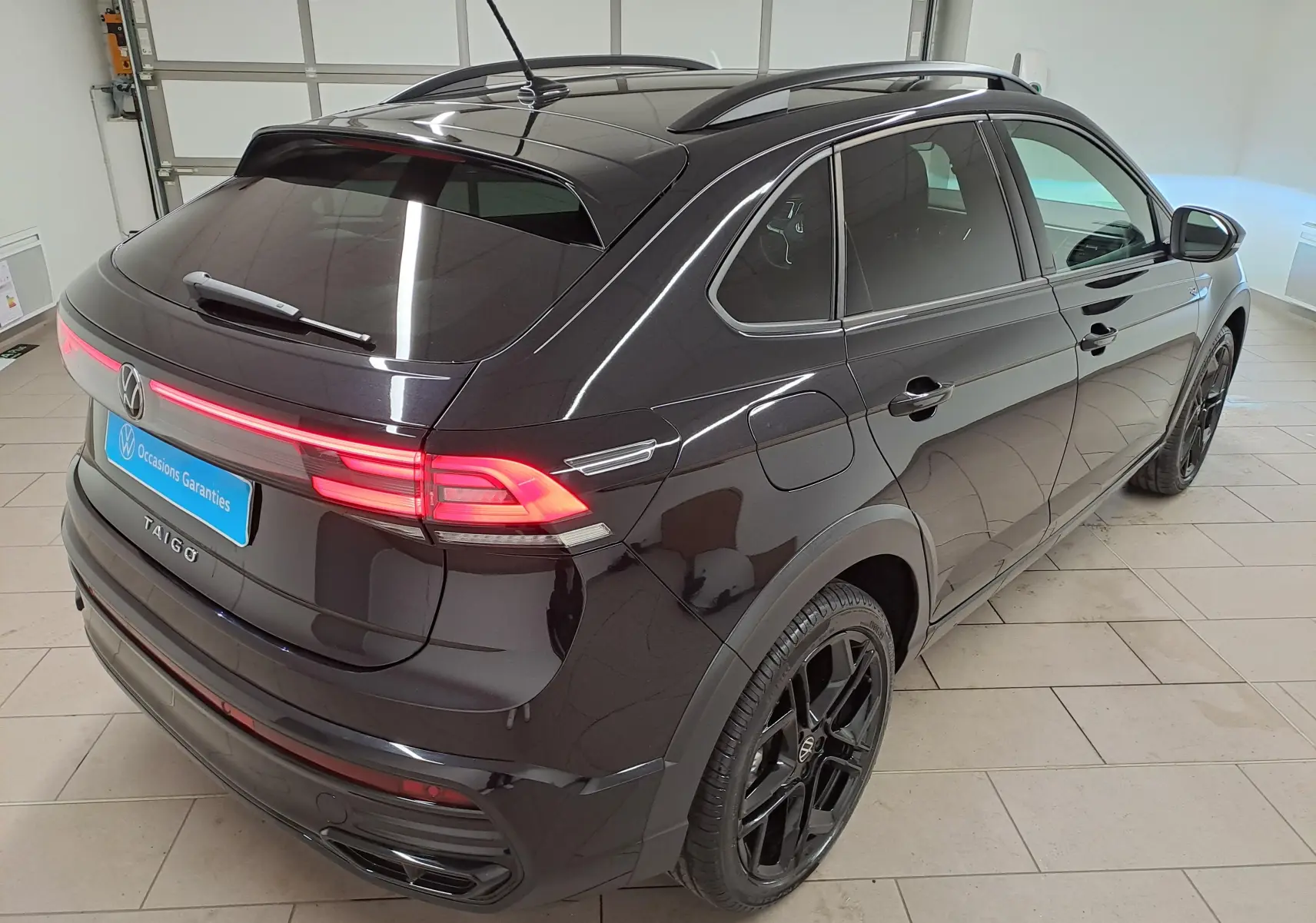 Vue 3/4 arrière droite du Volkswagen Taigo noir intense nacré avec jantes noires et feux arrière allumés en intérieur showroom.