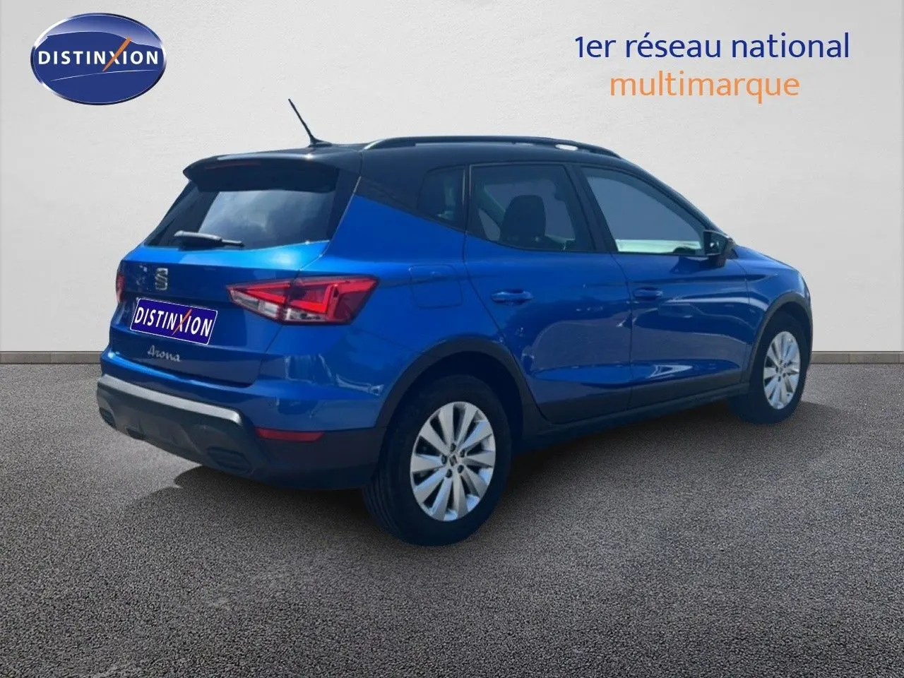 Vue 3/4 arrière droite d'une SEAT Arona bleu saphir métal avec toit noir, stationnée sur sol gris devant un mur blanc.