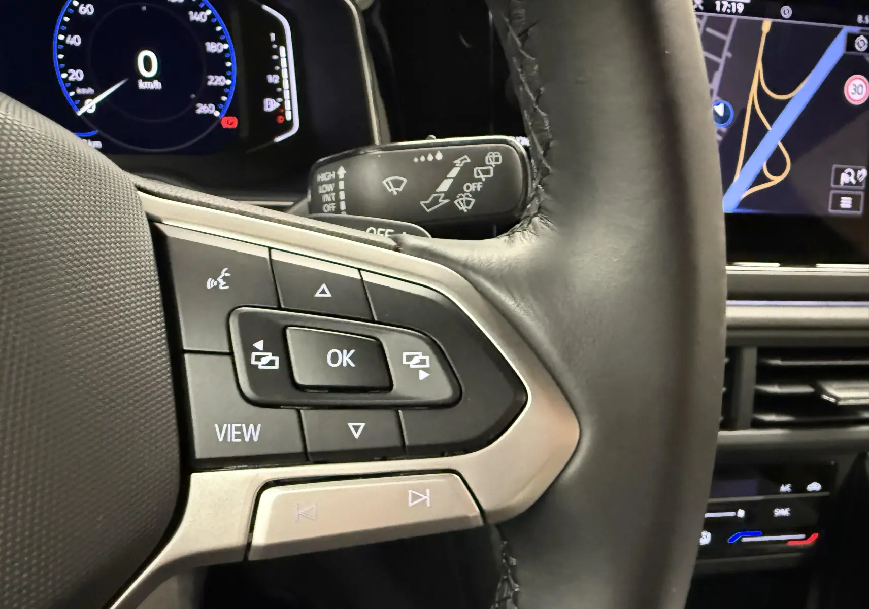 Gros plan sur les commandes du volant cuir noir du Volkswagen Taigo gris cendre, avec affichage digital et écran tactile en arrière-plan.