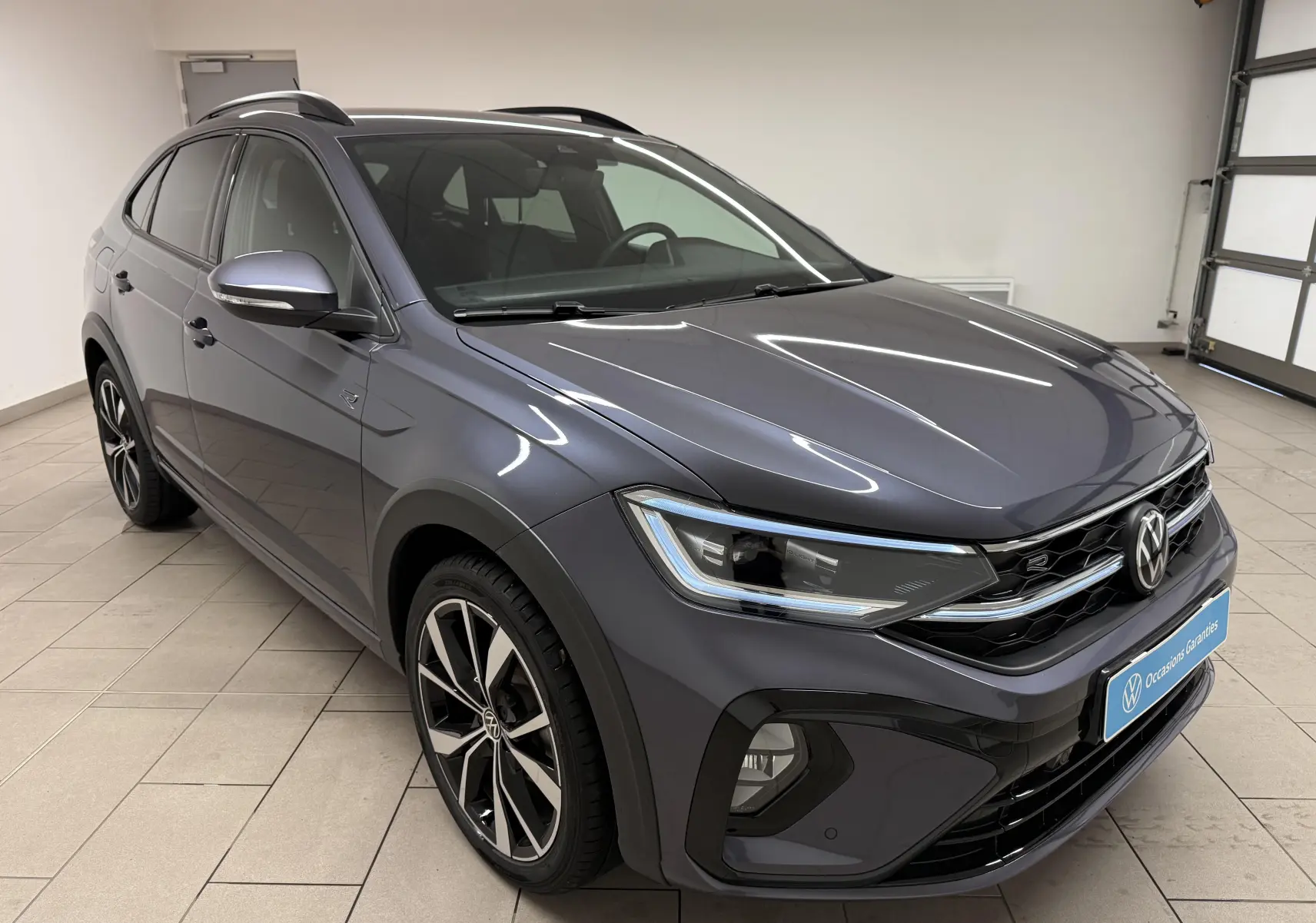 Volkswagen Taigo gris cendre en 3/4 avant droit, avec jantes alliage et calandre R-Line visible en intérieur showroom.