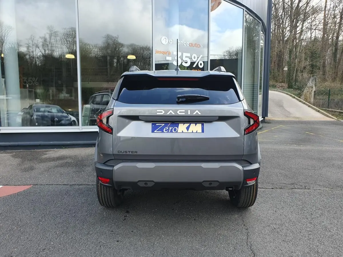 Vue arrière d'un Dacia Duster gris schiste 2026 avec feux LED et plaque d'immatriculation Zero KM devant un bâtiment vitré.