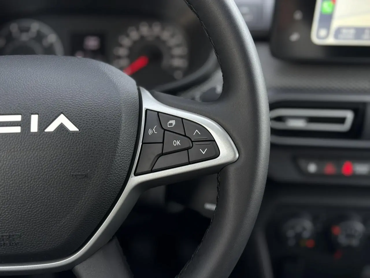 Gros plan sur le volant noir du Dacia Jogger 2024 avec commandes multifonctions et tableau de bord flou en arrière-plan.