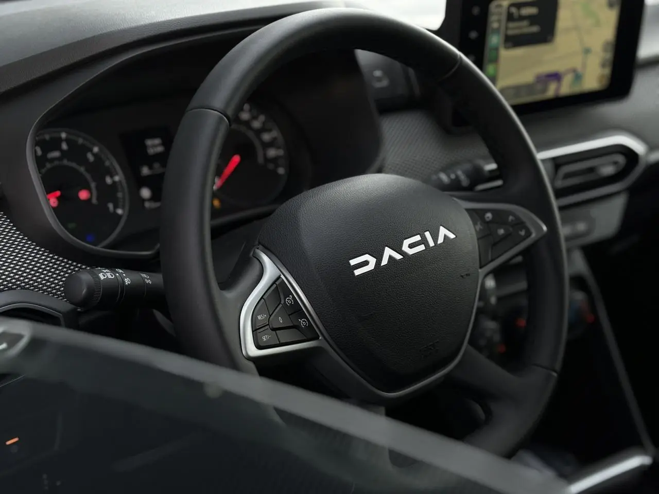 Vue rapprochée du volant noir de la Dacia Jogger 2024 avec commandes intégrées et écran GPS tactile visible en arrière-plan.