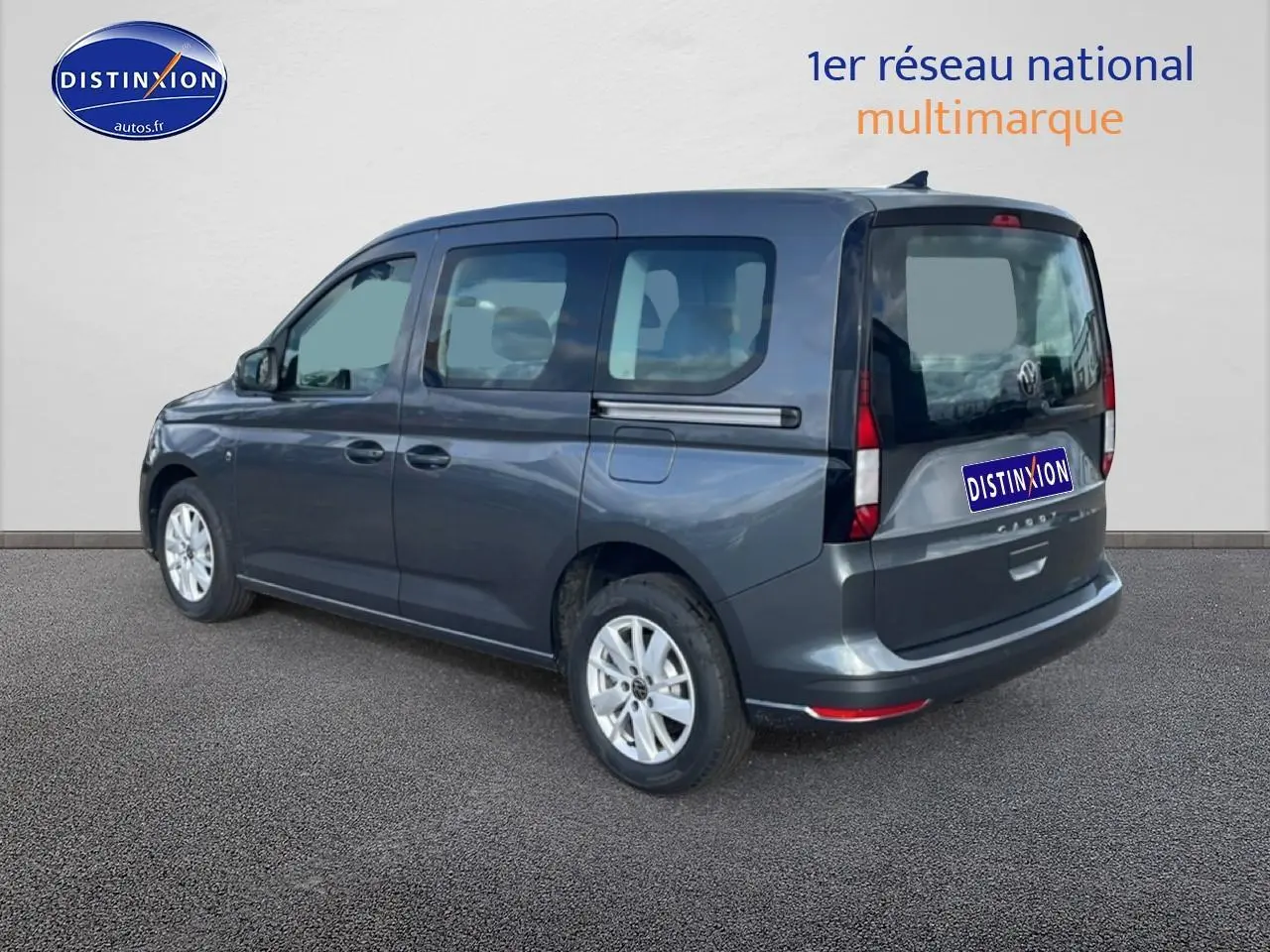 Volkswagen Caddy gris indien vu en 3/4 arrière droit avec vitres teintées et barres de toit visibles.