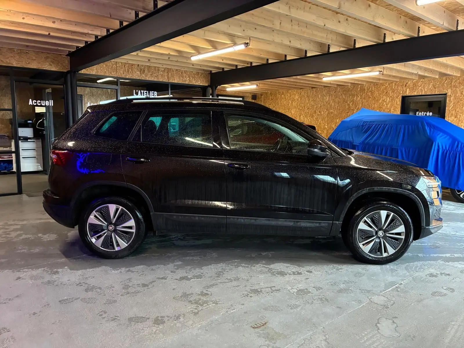 Vue de profil côté gauche d'un Skoda Karoq noir 2023 dans un garage avec toit en bois et sol béton.