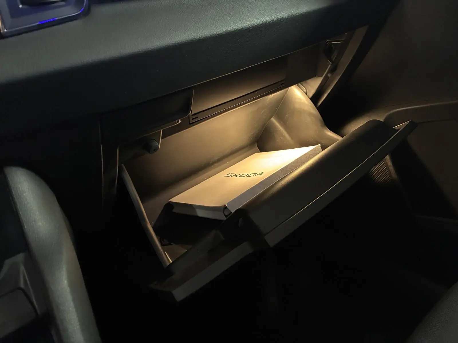 Vue intérieure en plongée sur la boîte à gants ouverte du Skoda Karoq noir, avec manuel éclairé à l'intérieur.