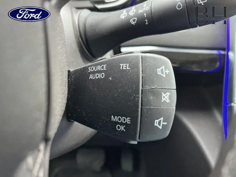 Gros plan sur la commande audio au volant noir, avec inscriptions blanches, dans un intérieur sombre.