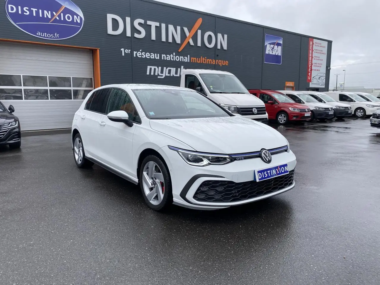 Volkswagen Golf 1.4 eHybrid blanche vue 3/4 avant avec calandre noire et jantes argentées sur parking humide