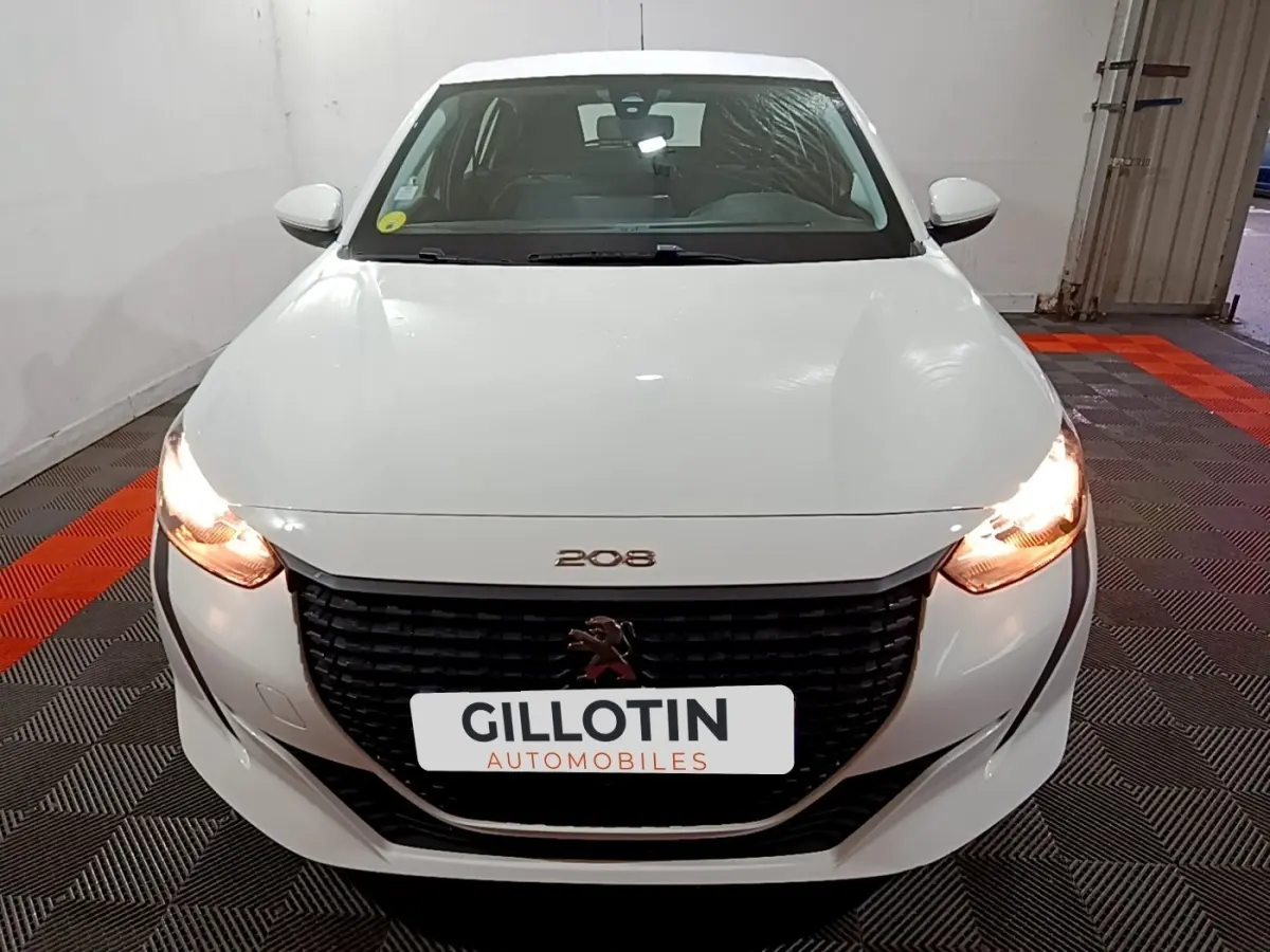 Vue frontale d'une Peugeot 208 blanche avec phares allumés et calandre noire distinctive dans un garage.