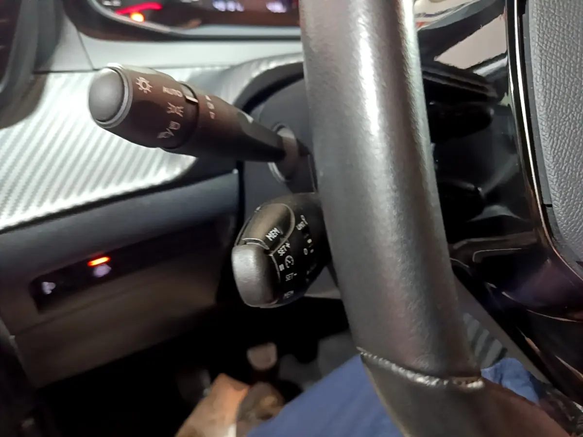 Gros plan sur la colonne de direction et les commandes derrière le volant d'une Peugeot 208 blanche, intérieur noir.
