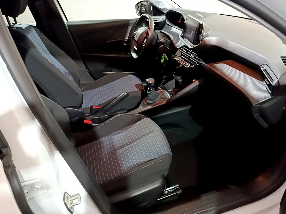 Vue intérieure côté conducteur de la Peugeot 208 blanche, montrant les sièges tissu à motifs et la console avec écran tactile.