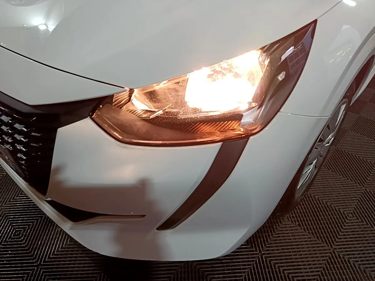 Gros plan sur le phare avant droit allumé d'une Peugeot 208 blanche, mettant en valeur la calandre et la jante.
