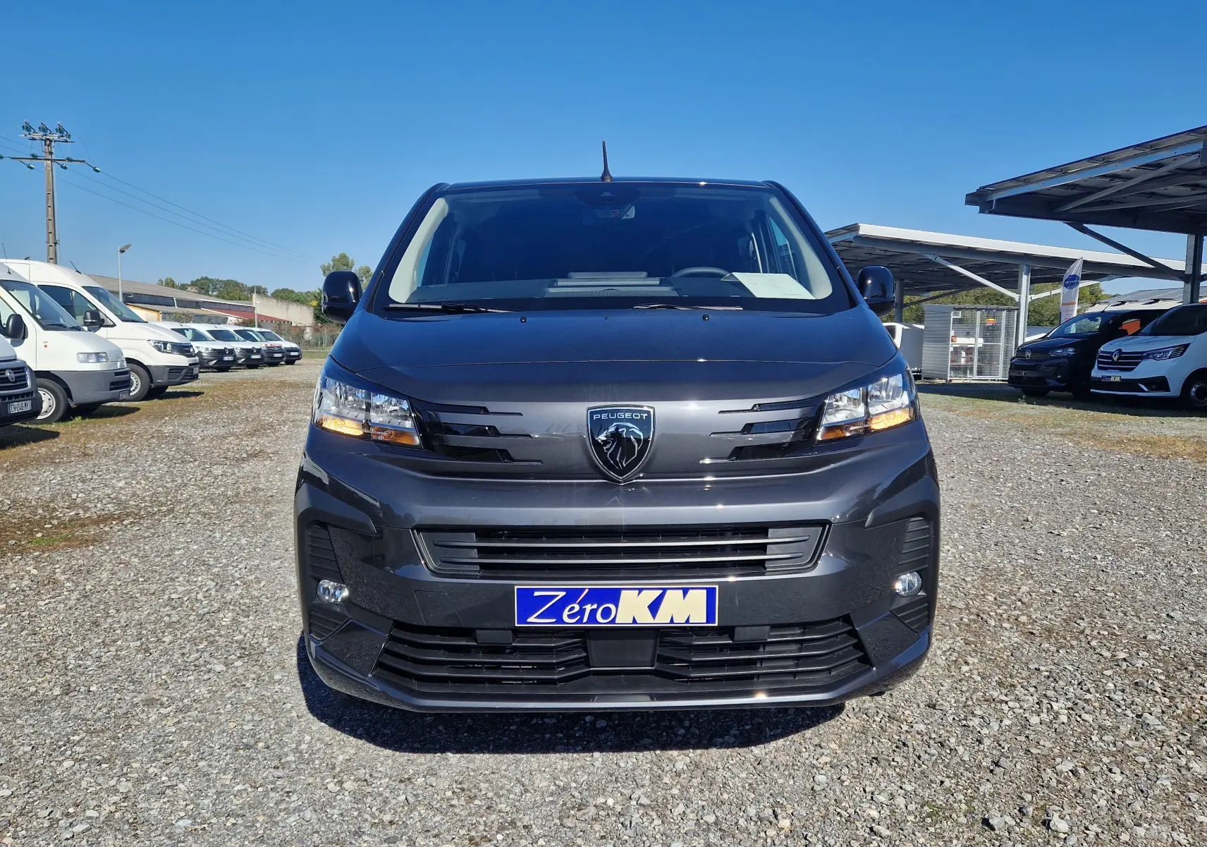 Vue frontale d'un Peugeot Expert Cabine Approfondie gris titane avec plaque Zéro KM sous un ciel bleu.