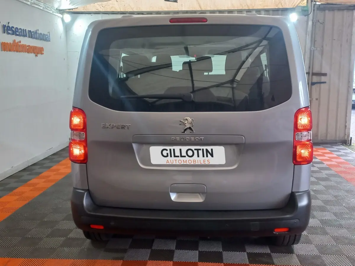 Vue arrière d’un Peugeot Traveller gris clair 2021 avec feux allumés et logo visible dans un garage.