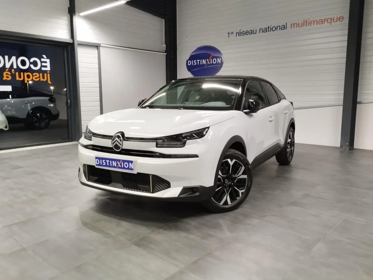 Citroën C4 2025 blanc Okenite avec toit noir, vue 3/4 avant en intérieur showroom, jantes alliage noires et chromées.