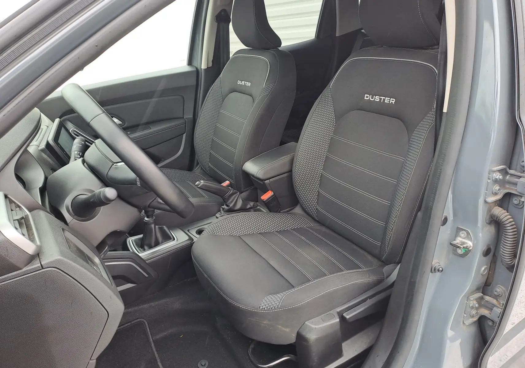 Vue intérieure côté conducteur du Dacia Duster gris urbain 2024, sièges noirs avec logo Duster brodé.