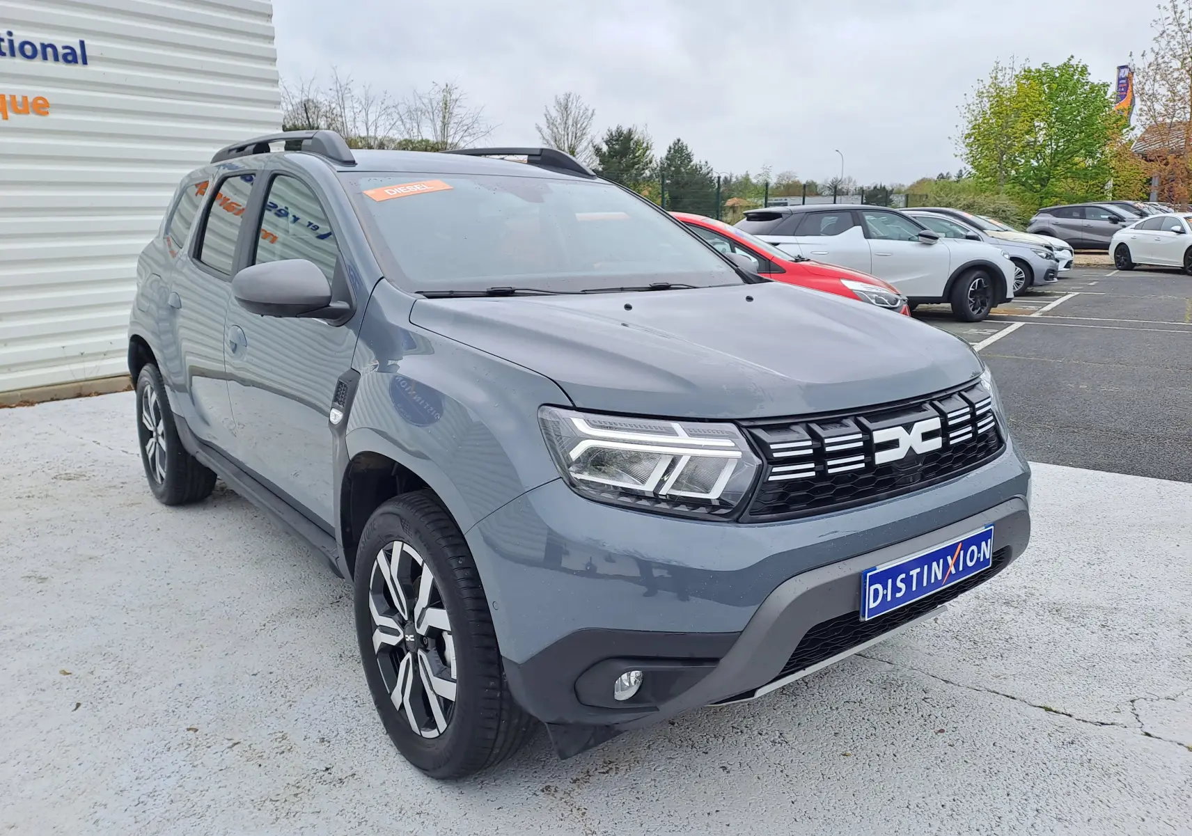 Dacia Duster gris urbain en 3/4 avant droit, avec calandre noire et jantes bi-ton visibles sur parking.