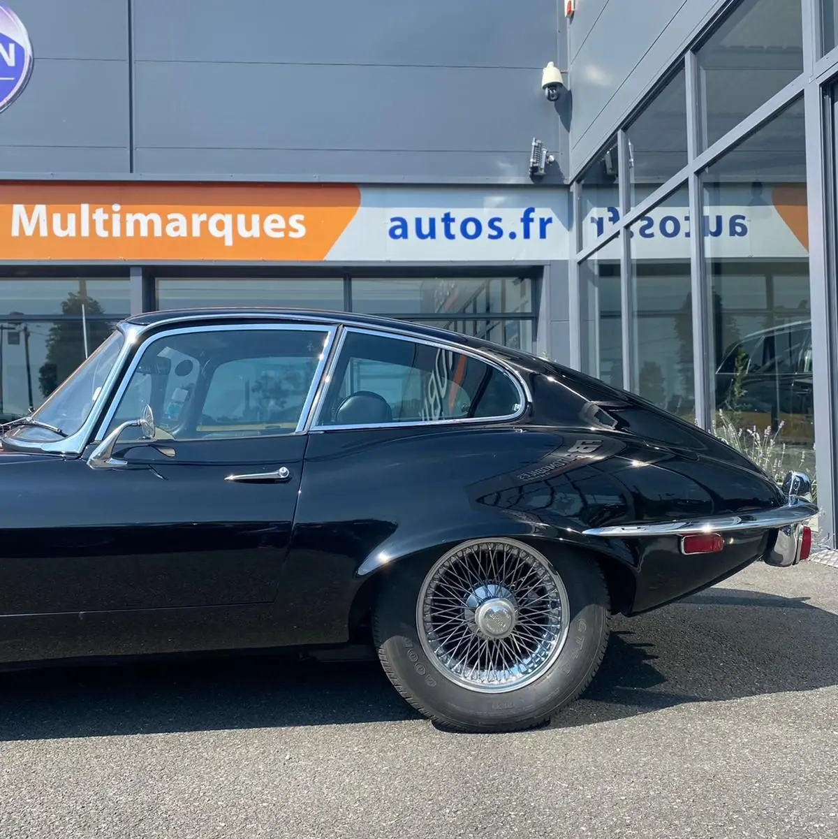 Profil côté gauche d'une Jaguar S-Type Type E Coupé noir de 1970 avec jantes à rayons chromés, garée devant un bâtiment moderne.