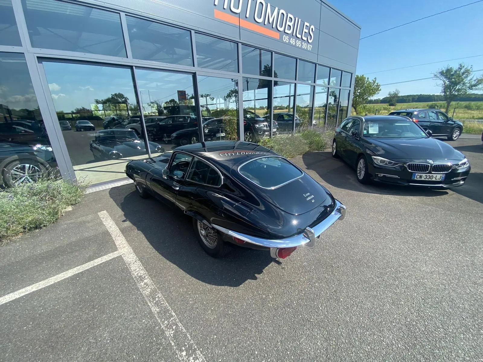Jaguar Type E Coupé noir vue 3/4 arrière droit, garée devant un showroom avec jantes à rayons chromés.