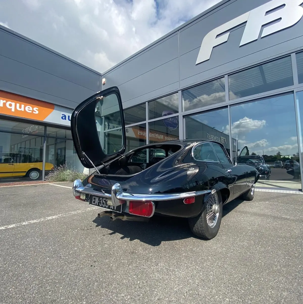 Vue 3/4 arrière gauche d'une Jaguar S-Type noire de 1970 avec coffre ouvert, garée devant un bâtiment moderne.