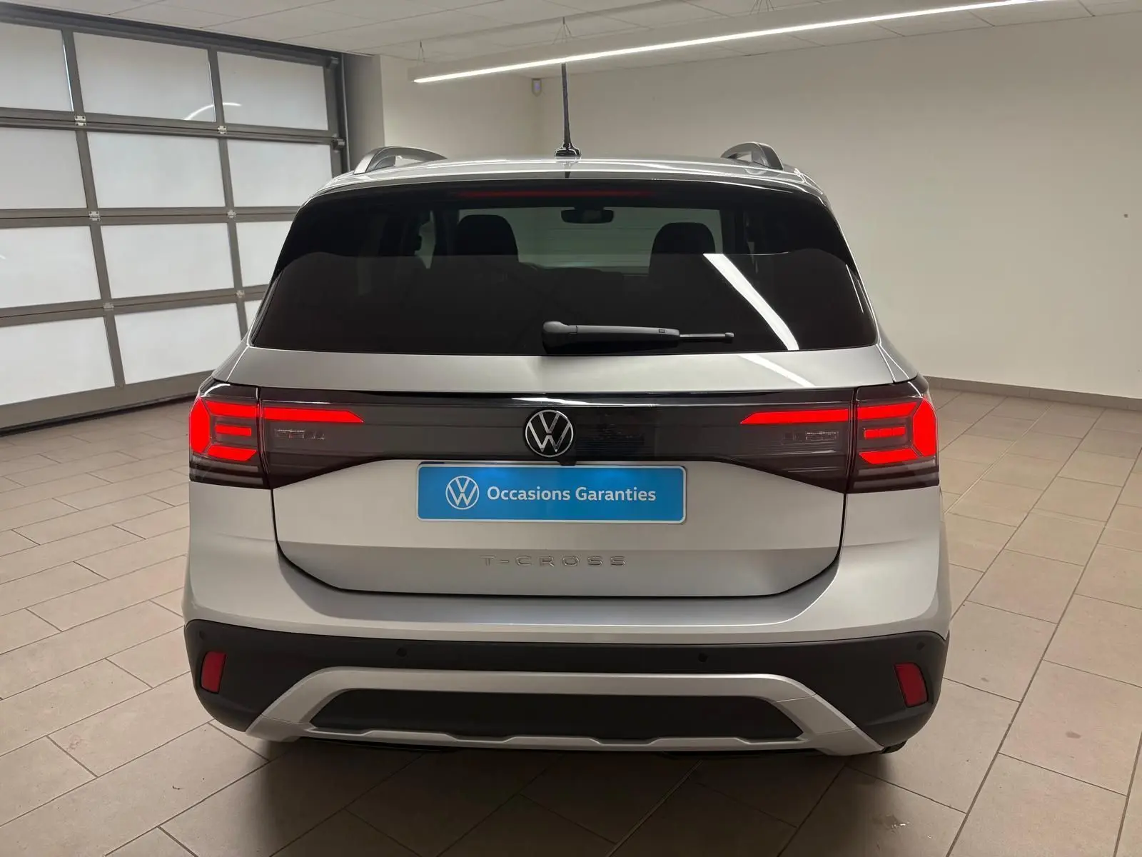 Vue arrière d'un Volkswagen T-Cross 2024 couleur reflet d'argent avec feux arrière LED allumés en intérieur.