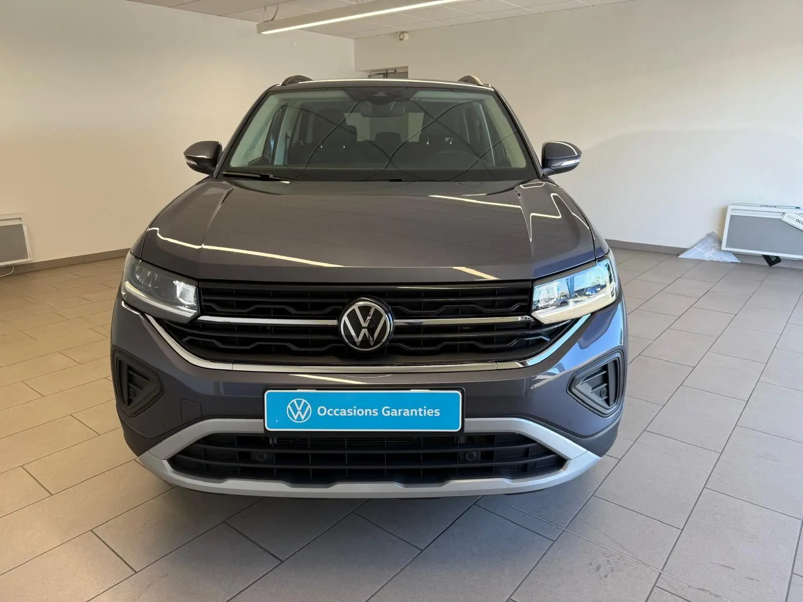 Vue avant d'un Volkswagen T-Cross 2025 gris cendre, avec calandre noire et logo VW centré, en intérieur showroom.